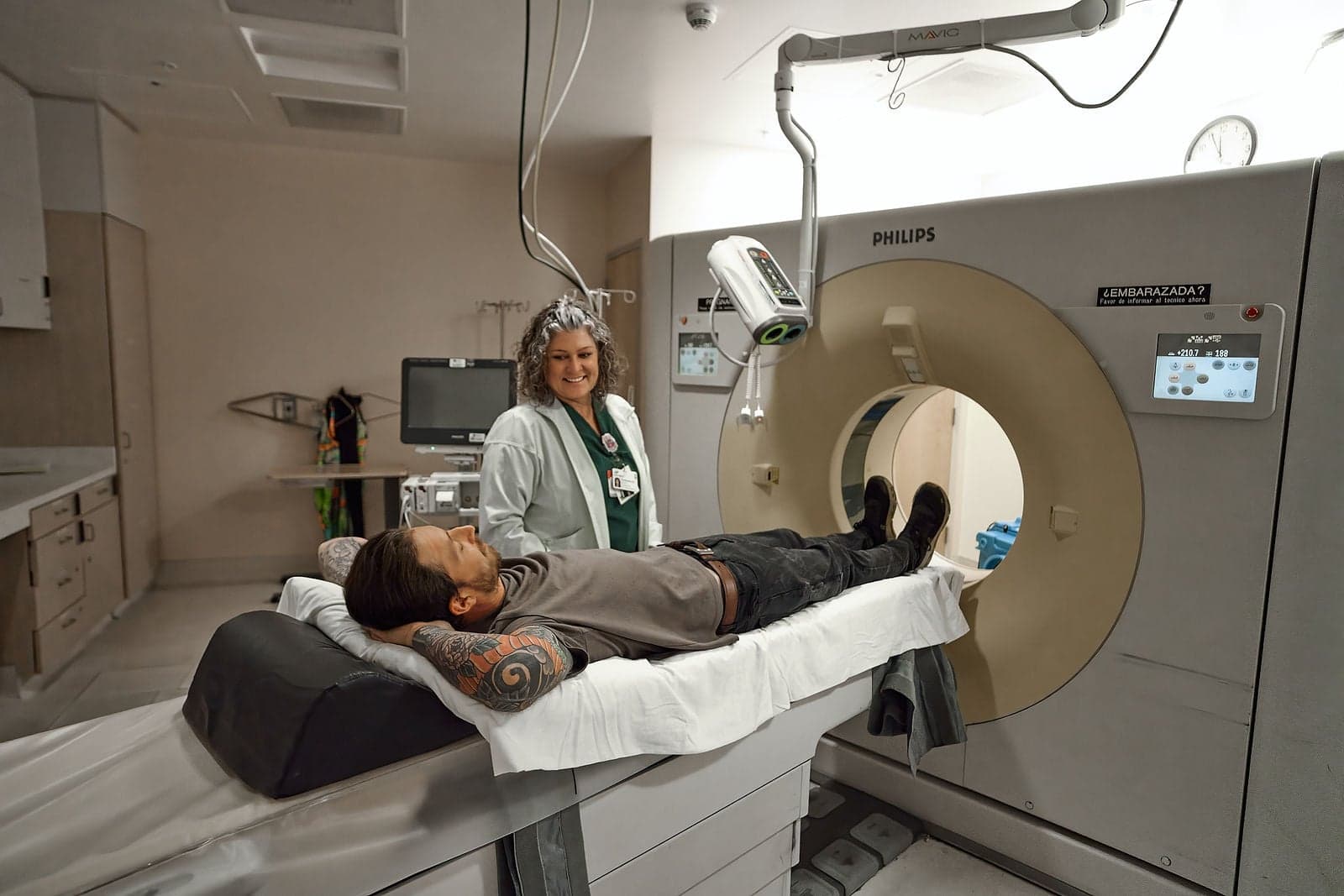 A technologist smiles beside a patient lying on the table of a CT scanner.