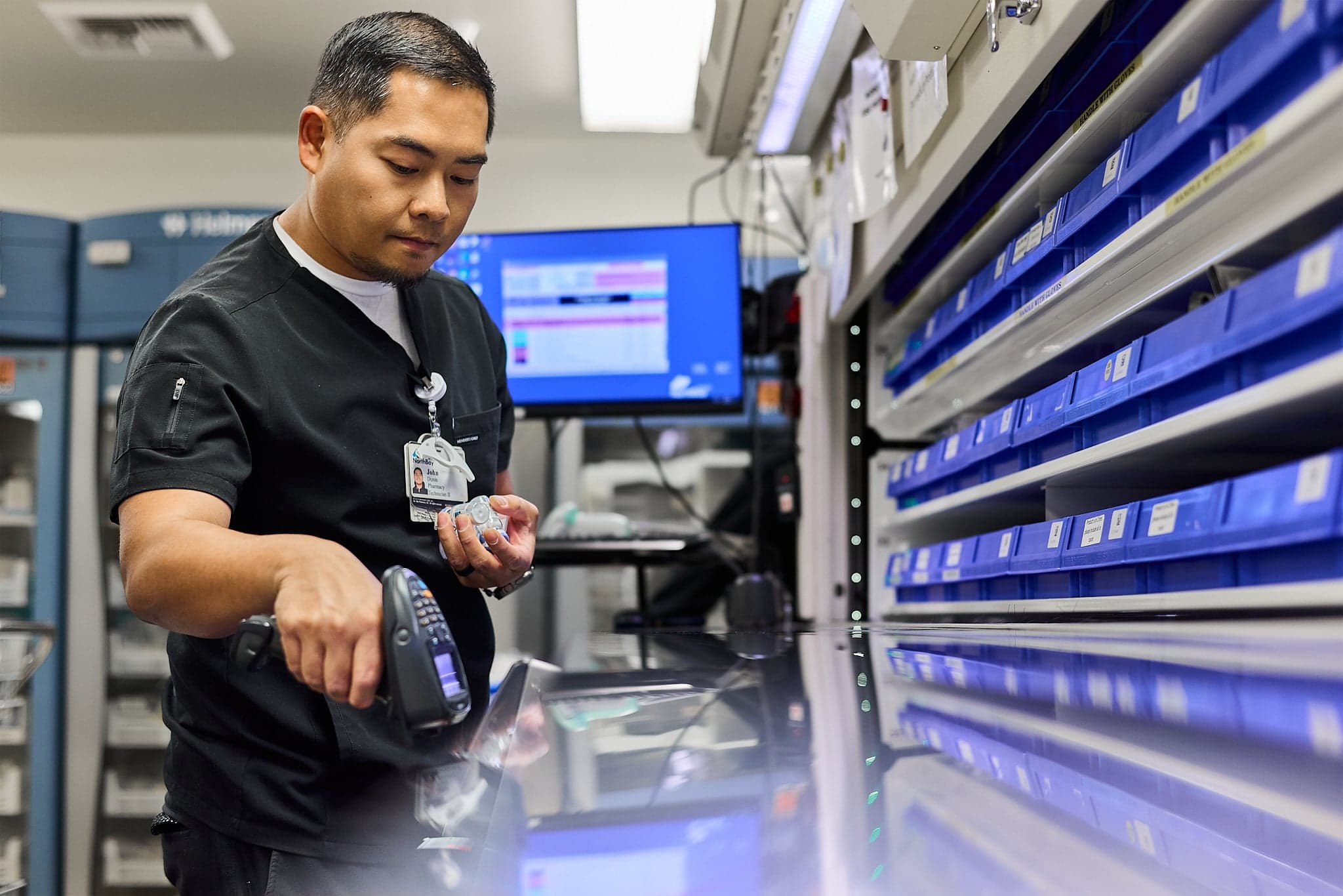 A pharmacy team member scans a medication in front of shelves of blue bins.