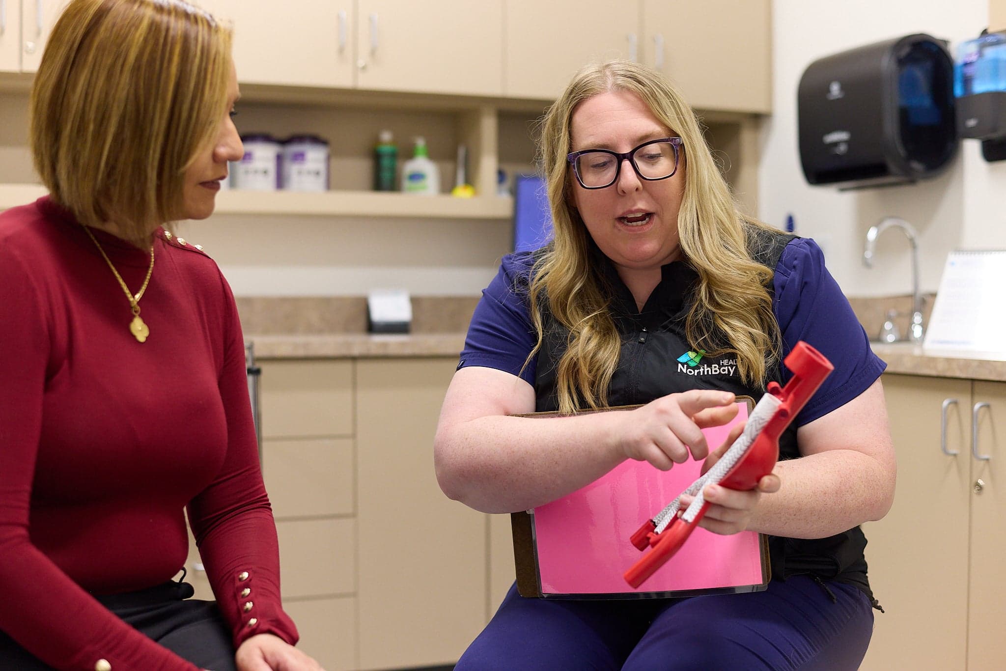 A Northbay Health team member holds a red breathing device and a clipboard while talking with a woman in an exam room.