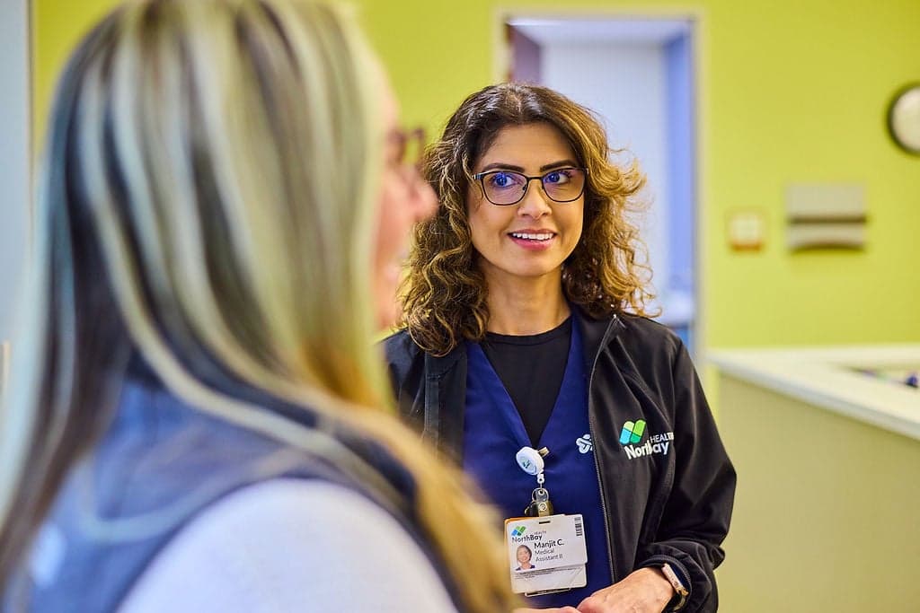 A smiling Northbay Health team member in glasses and scrubs talks with a woman in a clinic.