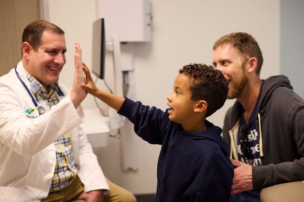 A doctor in a white coat high-fives a young boy sitting beside a smiling adult in an exam room.