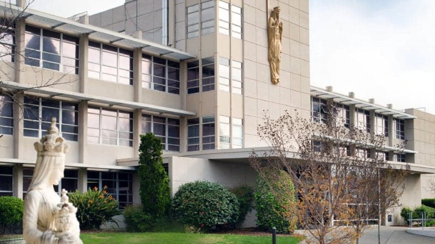 Modern institutional building with glass windows, white stone facade, and golden statue on corner, surrounded by manicured la