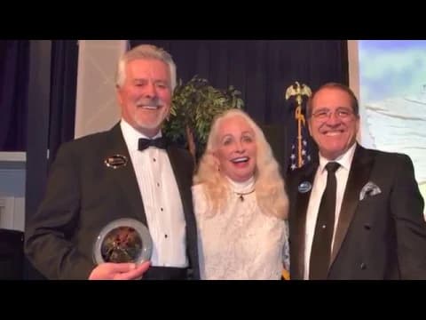 Three people in formal attire at an event, with two men in tuxedos holding awards flanking a woman in a white dress