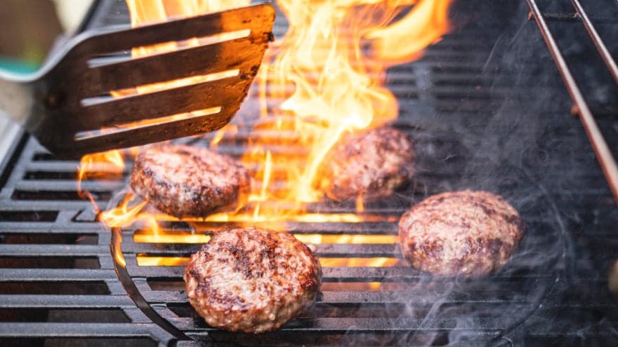 Close-up of four hamburgers being grilled with a large, fiery flare-up in the middle.