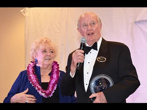 Man in tuxedo holding microphone and award standing next to woman in blue dress with lei at formal event
